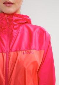Veste imperméable en rose vif et corail avec zip avant et capuche réglable. Présente un logo sur la zone de la poitrine.