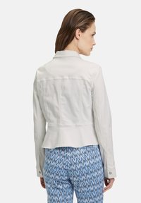 Veste en denim blanche avec un design structuré, bord peplum et poignets boutonnés, associée à un pantalon bleu à motifs présentant des formes de diamants.
