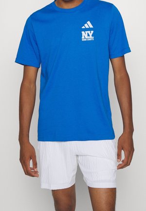 Mann trägt ein königsblaues Adidas T-Shirt mit dem Logo "NY Hard Courts" und weiße, strukturierte Shorts, steht vor einem einfarbigen Hintergrund.
