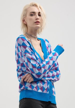 Blonde Person, die eine geometrisch gemusterte Strickjacke in Blau, Lila und Weiß mit großen Knöpfen und einer silbernen Kettenhalskette trägt.