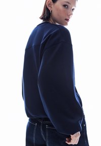 Laivastonsininen collegepaita rentolla istuvuudella, lasketuilla harteilla ja resoriremmuilla, yhdistettynä tummiin farkkuihin. Sileä tekstuuri, vähäiset yksityiskohdat.