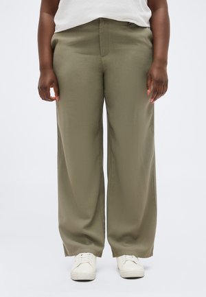 Person trägt olivgrüne Hosen mit weitem Bein, weiße Turnschuhe und ein weißes Hemd vor einem schlichten hellen Hintergrund.