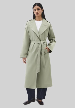 Kvinde iført en lysegrøn trenchcoat med bælte over en hvid top, mørke jeans og sorte sko, stående mod en ensfarvet grå baggrund.