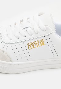 Baskets en cuir blanc avec détails perforés, lacets blancs et logo doré "Versace Jeans Couture" sur le côté.