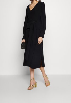 Robe noire portefeuille à manches longues, décolleté en V et taille nouée. Comprend des fentes latérales et est assortie à des sandales à talons bloc dorées avec des lanières.