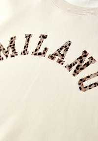 Sweatshirt beige avec le mot "MILAN" en police imprimée léopard marron. Texture lisse avec détail de col côtelé.