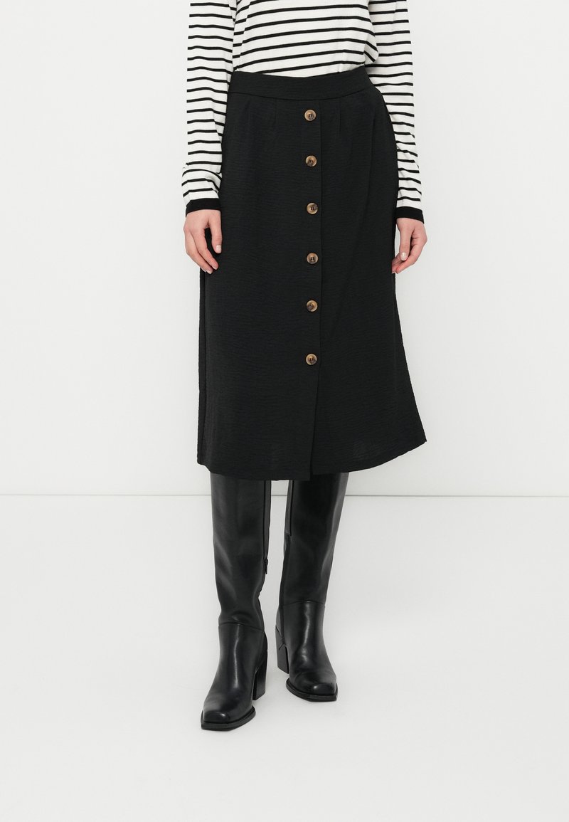 Μαύρη μίντι φούστα με κούμπωμα μπροστά με κουμπιά, υφή υφάσματος και ψηλή μέση. Φοριέται με μαύρες μπότες μέχρι το γόνατο και ριγέ μπλούζα με μακριά μανίκια.