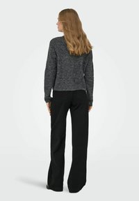 Kvinna med långt blont hår som bär en m�örkgrå tröja och svarta vida byxor, stående vänd bort från kameran mot en enkel bakgrund.