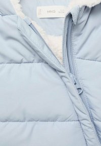 Jachetă puf galben deschis cu fermoar parțial deschis, arătând căptușeala din fleece alb și eticheta de îmbrăcăminte vizibilă în guler.