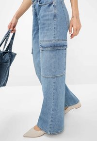 Helle blaue Denim-Cargohosen mit weitem Bein, aufgesetzten Taschen an den Oberschenkeln und kontrastierenden Nähten. Ein beiger Schuh ist unter dem Saum sichtbar.