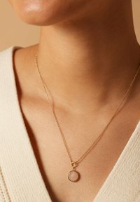 Collier doré avec un pendentif rond en pierre rose, suspendu à une chaîne délicate. Porté sur une clavicule nue.
