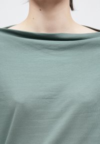 Svetlozelený, mäkký top z látky s uvoľneným výstrihom do loďky a hladkou textúrou, s jemným záhybom cez oblasť hrudníka.