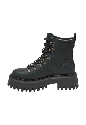 Personne sortant d’un véhicule portant des bottes noires épaisses à lacets, une jupe à carreaux et un trench-coat sombre sur un trottoir urbain ensoleillé.
