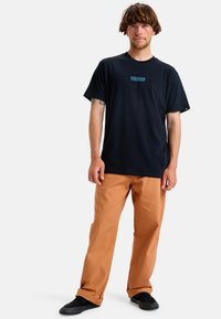 Svart bomullströja med blå text "FRIHET FRÅN KAOS", tillsammans med oranga byxor och svarta skor, i en avslappnad passform.