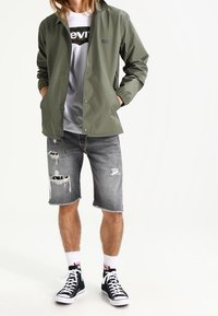 Persona con chaqueta verde de botones sobre una camiseta gráfica blanca, pantalones cortos de mezclilla gris desgastados, calcetines blancos y zapatillas altas negras.