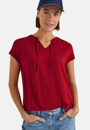 Femme portant un sweat à capuche rouge à manches courtes rentré dans un jean bleu taille haute avec des détails de boutons et une casquette de baseball bleu foncé.
