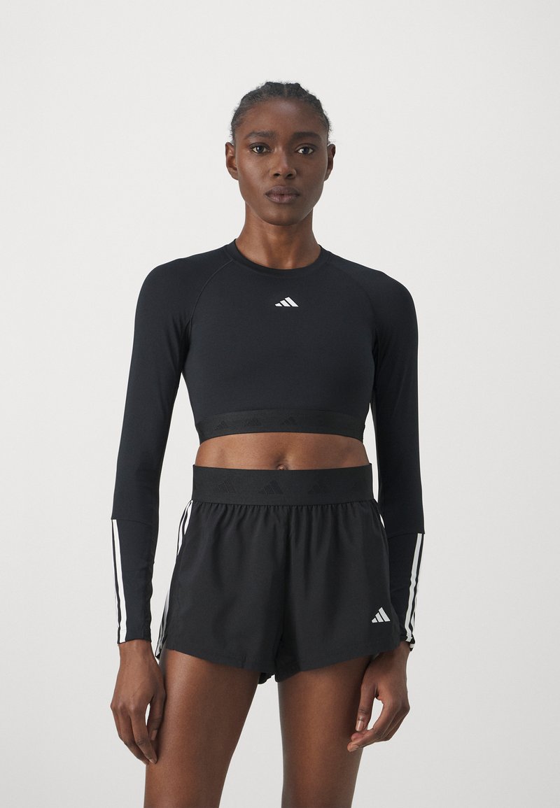 Crni atletski crop top s dugim rukavima i bijelim logotipom, uparen s crnim šortsama koje imaju širok pojas i bijele bočne pruge.
