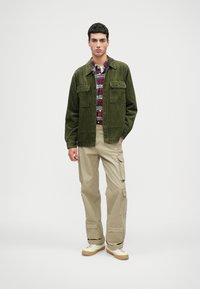 Πράσινο παντελόνι βελούδο πάνω από πουκάμισο με κόκκινο και μαύρο καρό, συνδυασμένο με μπεζ cargo παντελόνι και λευκά sneakers, με λειτουργικές τσέπες.