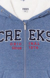 Sudadera azul con cremallera y forro de felpa, con letras bordadas texturizadas en azul marino y rojo, y bolsillos frontales. La etiqueta dice "CREEKS Desde 1974."