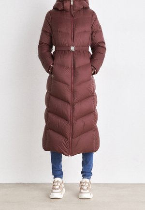 Długa bordowa pikowana puchowa kurtka z kapturem i paskiem, noszona z niebieskimi dżinsami i beżowo-białymi trampkami, stojąca na jednolitym tle.