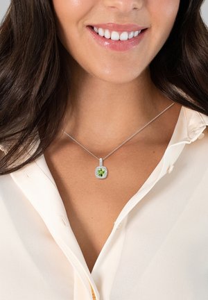 Collier en argent avec un pendentif rond en pierre précieuse verte entouré de petites touches blanches. Porté par-dessus une chemise de couleur claire.