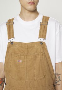 Homme portant une salopette beige avec une poche avant et un t-shirt blanc, des tatouages colorés visibles sur les deux avant-bras.