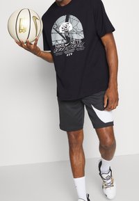 Camiseta negra de Nike con un gráfico de baloncesto y texto, combinada con pantalones cortos oscuros que tienen un acento blanco, sosteniendo una pelota de baloncesto color crema.