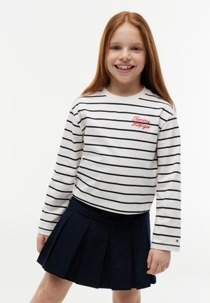 Smilende pige med langt rødt hår iført en hvid og marineblå stribet langærmet skjorte og et marineblåt plisseret skørt, stående mod en hvid baggrund.