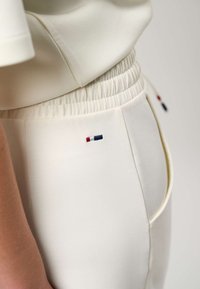 Približni posnetek osebe, ki nosi hlače z elastičnim pasom v barvi slonokosti, z majhno rdečo, belo in modro pravokotno oznako blizu roba žepa.