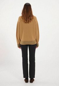 Pull en tricot beige avec des poignets en côtes et un ourlet inférieur, associé à un pantalon à rayures sombres. Le modèle est montré de dos.