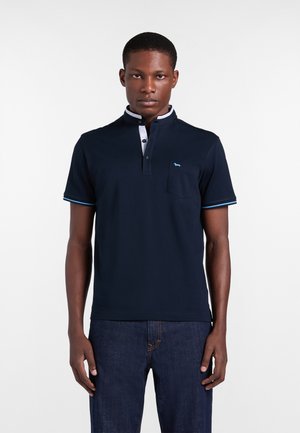 Uomo che indossa una polo blu navy a maniche corte con rifiniture bianche su collo e bordi delle maniche, abbinata a jeans blu scuro, in piedi davanti a uno sfondo semplice.