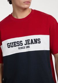Punane, valge ja tumesinine värviga blokeeritud T-särk ümarate kaelustega, millel on rinnaosas tumesinise värviga broderie "GUESS JEANS SINCE 1981".