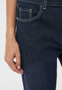 Jeans di denim blu scuro con una ripresa ravvicinata di una mano che tiene la cucitura laterale. Presentano cinque tasche e cuciture a contrasto. Texture liscia.