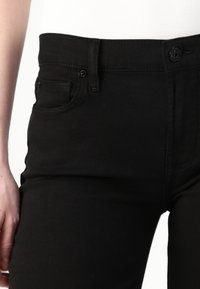 Črne denim hlače z strukturiranim pasom, sprednjimi žepi in gumbom za zapiranje. Material se zdi gladek z blagim raztezanjem.