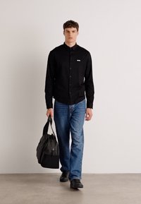 Black button-up shirt, blue jeans, and black shoes. Model carries a black bag. Plain design, standard fit, smooth fabric, minimal branding.