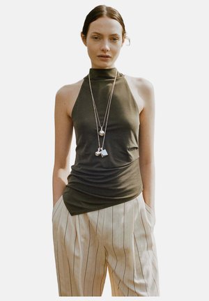 Femme avec un haut sans manches foncé, pantalon beige à rayures, mains dans les poches, portant deux longs colliers argentés avec des pendentifs géométriques.