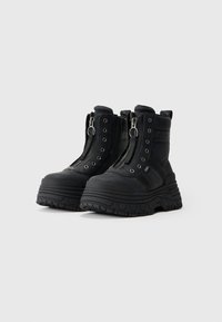 Un par de botas chunky negras con plataformas, con cremalleras frontales, ojales de metal y suelas texturizadas sobre un fondo claro y liso.