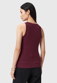 Mujer con cabello largo y negro que lleva una blusa sin mangas color burdeos y jeans negros, de pie de espaldas contra un fondo claro y liso.