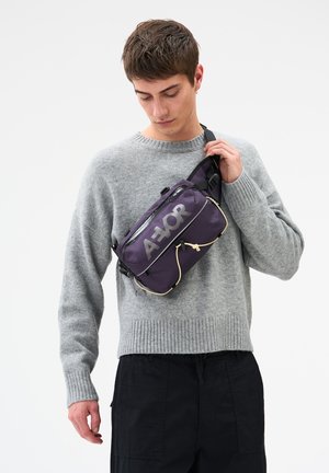 Junger Mann in grauem Pullover und schwarzer Hose, der eine lila Umhängetasche mit Gurt und auffälligem weißem Logo justiert.