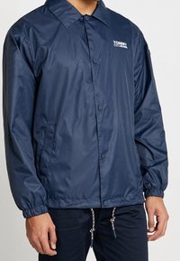 Chaqueta ligera de color azul marino con cuello puntiagudo, cierre de botones, puños elásticos y bolsillos laterales. Presenta el logotipo "TOMMY JEANS" en blanco.