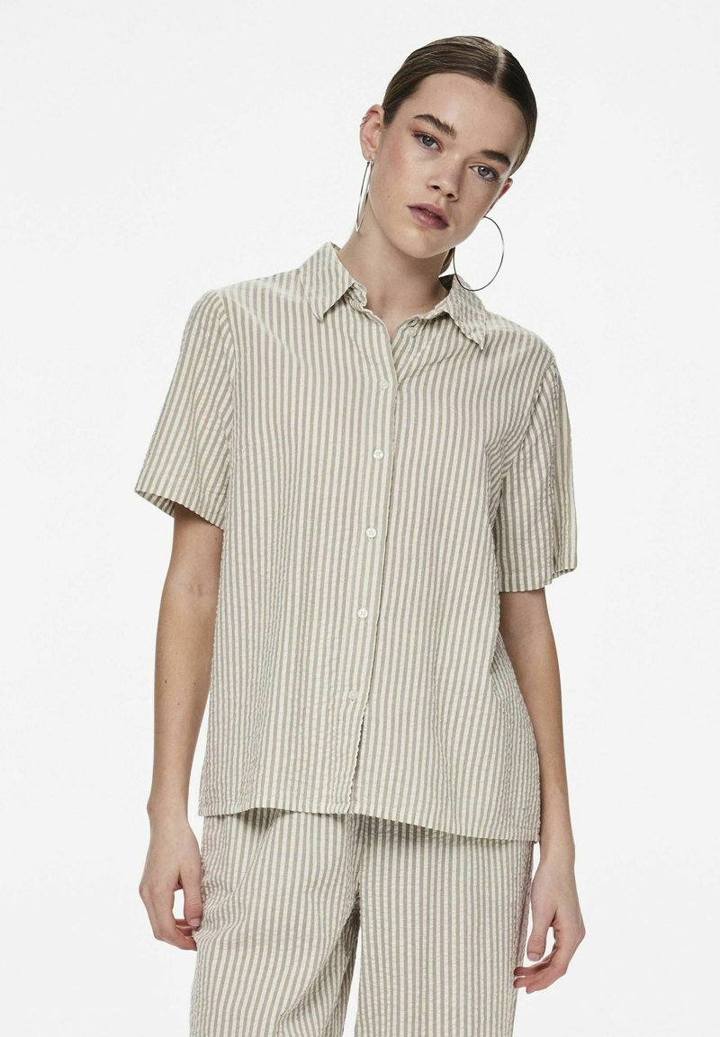 Camicia a maniche corte con bottoni di un beige chiaro a righe verticali. Realizzata in un tessuto testurizzato, presenta un colletto classico e una vestibilità comoda.