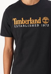 Černé bavlněné tričko s vyšitým logem "Timberland" v žluté a bílé barvě, s nápisem "Established 1973" a emblémem stromu.
