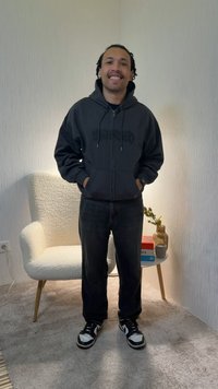 Young man smiling with hands in pockets, wearing black hoodie, pants, and sneakers, standing near white armchair and side table with books and vase.