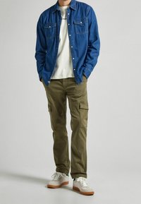 Blå denimskjorta med knappar, vit t-shirt under, olivgröna cargobyxor och vita sneakers med bruna sulor. Avslappnad, lager-på-lager outfit.