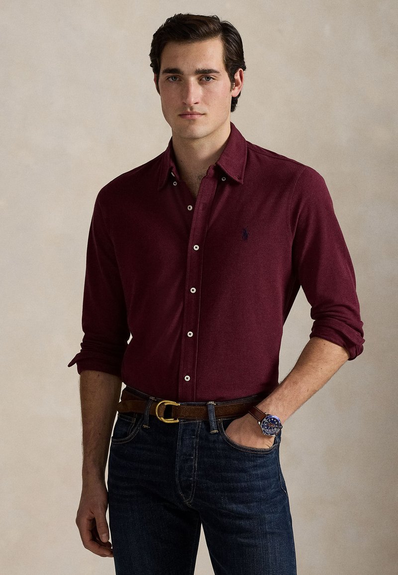 Joven con una camisa burdeos abotonada con las mangas remangadas, vaqueros azul oscuro, cinturón marrón y un reloj en su muñeca izquierda.
