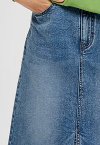 Hand in der Tasche eines blauen Jeansrocks, der mit einem grünen Oberteil getragen wird, deutet auf die genähte Naht und die Stoffstruktur hin.