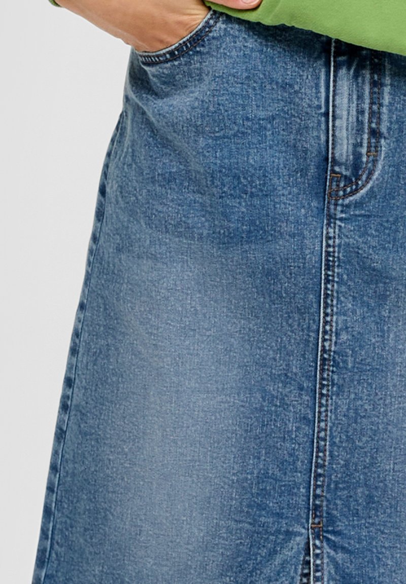 Hand in der Tasche eines blauen Jeansrocks, der mit einem grünen Oberteil getragen wird, deutet auf die genähte Naht und die Stoffstruktur hin.