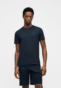 Άνδρας που φοράει σκούρο ναυτικό μπλε κοντομάνικο μπλουζάκι και αντίστοιχα σορτς, στέκεται μπροστά σε απλό γκρι ανοιχτό φόντο.