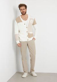 Cardigan crema con accenti beige, motivi a maglia testurizzati e bottoni in legno. Abbinato a pantaloni beige chiaro e sneakers bianche.