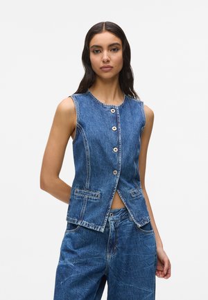 Jonge vrouw met een mouwloos blauw denim gilet met knopen en losse blauwe denim jeans, staand tegen een effen achtergrond.